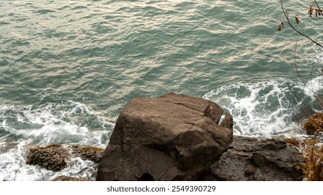 Close-up of a coastal rock formation with waves gently crashing against it, creating frothy white foam. The greenish-blue seawater ripples under soft natural light. - Powered by Shutterstock