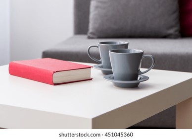 Close-up Of A Coffee Table With Two Grey Cups And A Book In Red Cover, Next To A Blurred Grey Sofa In The Background