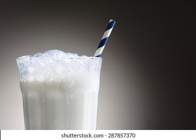 Closeup Of A Cold Glass Of Milk With A Blue And White Striped Drinking Straw. The Milk Glass Has A Frothy Top With Bubbles Running Down The Outside. Horizontal Over A Light Ot Dark Gray Background.