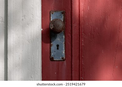 A closeup of a colorful antique red painted exterior wooden door with a rusty metal door knob and steel plate. There's a keyhole in the plate. The trim on the building has wide white weathered boards. - Powered by Shutterstock