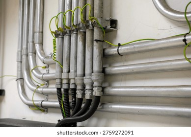 Close-up of a complex electrical conduit system, featuring metal pipes, junction boxes, and green grounding wires attached to a white wall.