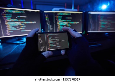 Close-up of computer hacker in black gloves holding mobile phone and taking a photo of computer software on it in dark office - Shutterstock ID 1557298517