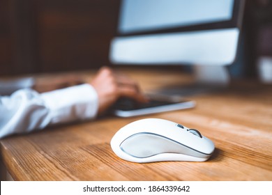 Close-up of a computer mouse on a table against the background of a working girl at a computer dressed in a white shirt. Selected focus on computer mouse on the wooden table. 