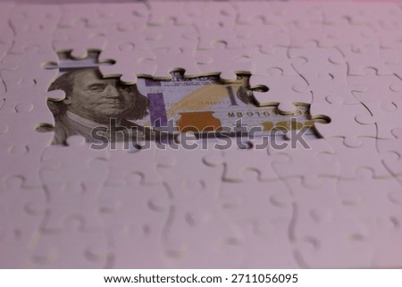 A closeup concept image of U.S. dollar bills partially revealed beneath an open white puzzle, symbolizing hidden money, financial strategy, savings, and economic insight.