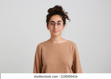 Closeup Of Confident Pretty Young Woman Student With Bun Of Dark Curly Hair Wears Beige Pullover And Glasses Feels Peaceful And Looks Directly In Camera Isolated Over White Wall Looking Serious  Calm