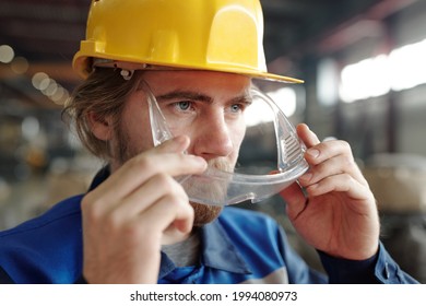 Close-up Of Confident Young Bearded Factory Worker In Yellow Hardhat Putting Safety Goggles On Face While Preparing For Work