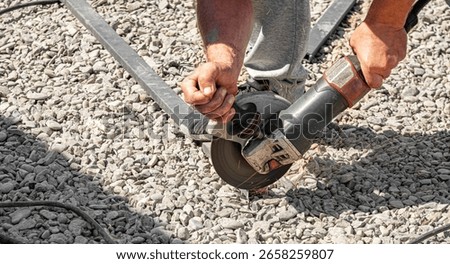 Close-up of a construction worker using an angle grinder to cut metal on a gravel surface, with visible sparks and intense manual effort.