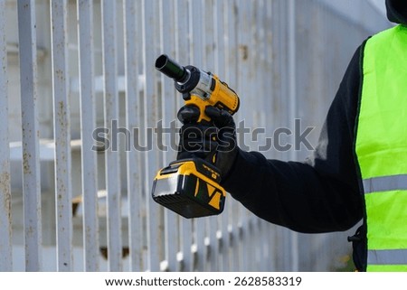 Close-up of a construction worker using a cordless impact drill to attach a metal fence. Protective gloves and a high-visibility vest for safety