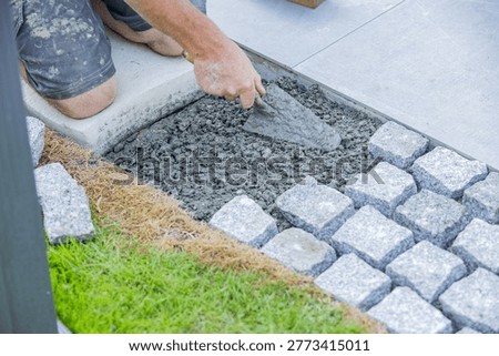 Close-up of a construction worker using a trowel while installing cobblestone pavement on a prepared base, showing landscaping and masonry work in progress.