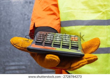 Close-up of a construction worker wearing protective gloves and safety vest holding a calculator. Concept of cost estimation, budgeting, and financial planning in building and renovation projects