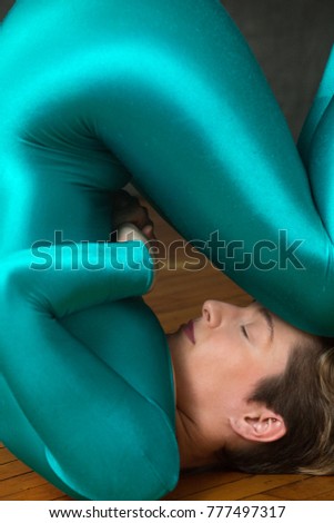 Closeup of a contemporary young woman dancer with short hair, wearing a blue unitard body suit, lying on her back on the wood floor of the studio with her knees on her forhead.