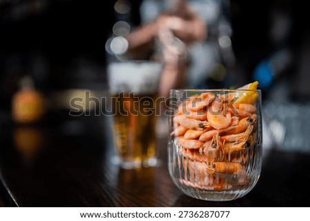 A close-up of cooked, shell-on prawns served with a lemon wedge in a glass on a dark wooden bar, with a blurry pint of beer in the background.