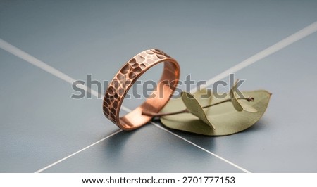 A close-up of a copper ring with a textured surface, resting beside a delicate leaf decoration on a sleek, modern surface.
