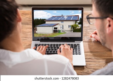 Close-up Of A Couple Searching For House With Solar Panel On Laptop Over Wooden Desk