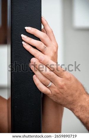 Close-up of couple's hands touching through black metal beam, showing gold engagement ring and silver wedding band, symbolizing connection, love, and commitment.