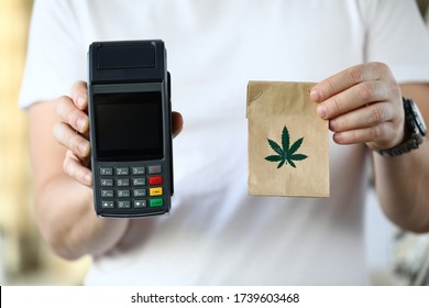 Close-up of courier holding paper package with traditional meds. Cannabis in bag with leaf symbol. Cashier machine for fast payment. Nfc technology counter and remedy concept