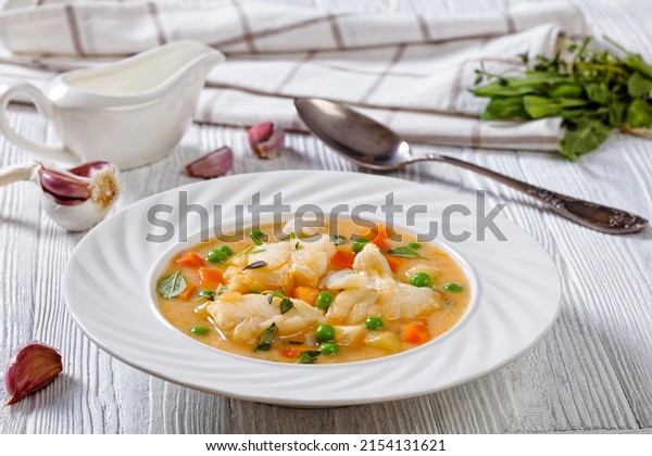close-up of Creamy fish chowder with cod,\
green peas, carrots, potatoes and plant based milk sprinkled with\
fresh sage, thyme in white bowl on white\
table