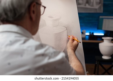 Closeup Of Creative Older Man Hand Holding Pencil Drawing Contour On Paper Canvas On Easel In Home Studio. Detail Of Elderly Artist Freehand Sketching Plaster Vase Model In Evening Art Workshop.