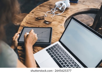 Close-up of creative woman sketching abstract graph on digital tablet, combining artistic expression and data visualization next to laptop and coffee in relaxed workspace