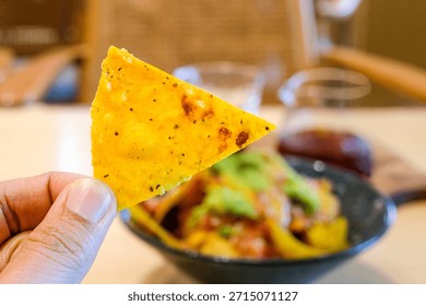 Close-up of a crispy nacho chip held by hand with blurred loaded nachos in the background on a cafe table. Perfect for food blogs, snack promotions, social media content, and casual dining concepts