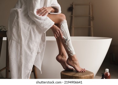 Closeup cropped image of female leg in shaving foam being shaved by woman in bathrobe, holding razor, making depilation in luxury bathroom near ceramic bathtub. Beauty procedures