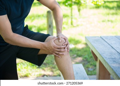 Closeup Cropped Portrait, Man In Black Shirt And Shorts Holding Knee In Severe Pain, Isolated Trees And Picnic Bench Outside Background. 