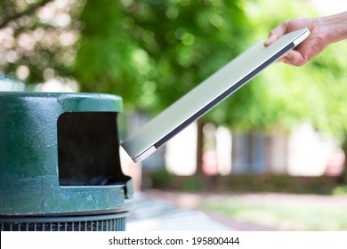 Closeup Cropped Portrait Of Someone Tossing Old Notebook Computer In Trash Can, Isolated Outdoors Green Trees Background