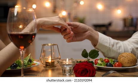 closeup with cropped shot of lovers holding hands over romantic valentine’s day dinner table with wine and red rose. the man gently touches the woman’s fingers.