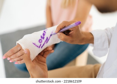 Close-up Cropped Shot Of Unrecognizable Male Funny Doctor Writing Joking Inscription On Cast Of Woman With Broken Hand Wrapped In Plaster Bandage. Concept Of Healthcare, Friendship And Fun.