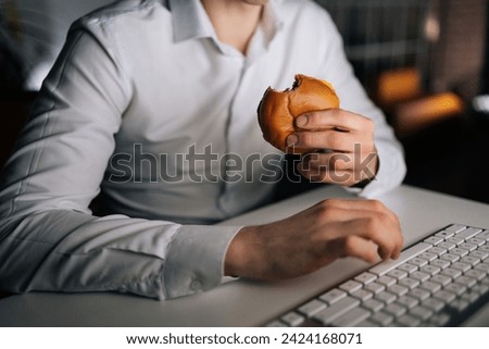 Close-up cropped shot of unrecognizable man eating burger working on computer sitting on desk at office workplace late night in dark room. Businessman eating fast food at laptop and reading email.