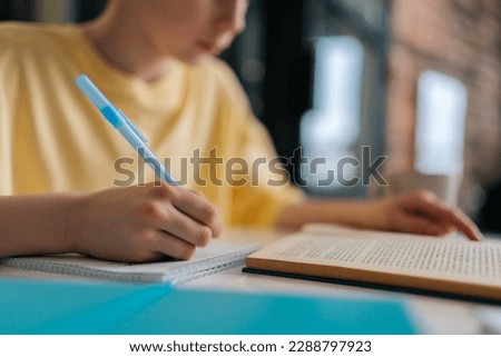 Close-up cropped shot of unrecognizable pupil boy studying at home writing in exercise book doing homework, learning sitting at table. Selective focus of schoolboy doing homework, reading textbook.