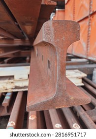 Close-up cross section view of a rusty steel railway track profile stacked in an industrial storage yard. Detailed texture of weathered iron rail track with pre-drilled joint holes.