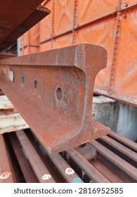 Close-up cross section view of a rusty steel railway track profile stacked in an industrial storage yard. Detailed texture of weathered iron rail track with pre-drilled joint holes.