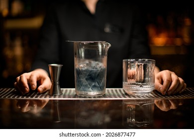 Close-up of crystal mixing cup with cold blue cocktail and old-fashioned glass and steel jigger stand side by side on the bar. Hands of woman bartender on bar counter