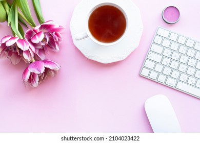 Closeup cup of black tea with pink lip balm, wireless white keyboard and mouse, three peony tulips on pink copyspace