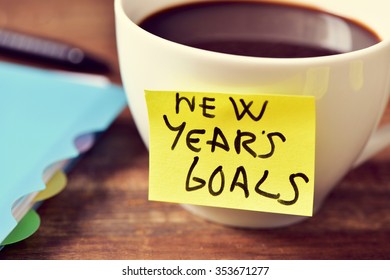 Closeup Of A Cup Of Coffee With A Yellow Sticky Note With The Text New Years Goals Written In It And A Notebook And A Pen On A Rustic Wooden Table