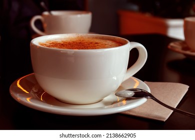 Close-up of cup of turkish traditional winter hot drink salep with cinnamon. White ceramic mug on plate and little spoon on napkin. Blurred background. Selective focus.