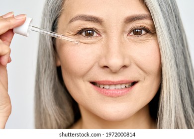 Closeup cut portrait of senior mature older Asian woman touching clean face eye contour with antiaging pipette serum essence oil looking at camera. Anti wrinkle prevention skin care products concept.