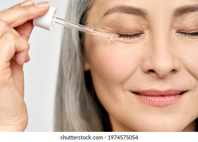 Closeup Cut Portrait Of Senior Mature Older Asian Woman With Closed Eyes Touching Face Eye Contour With Antiaging Pipette Serum Essence Oil. Anti Wrinkle Prevention Eye Skin Care Products Concept.