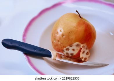 Close-up Of A Cut Rotten Apple On A Plate With A Knife. Symmetrical Spots Of Decay On The Cut Of The Fruit. Selective Focus. The Concept Of Skin Diseases.