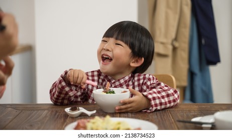 Closeup Of A Cute Asian Little Kids Boy Holding Spoon And Bowl And Looking At His Family With A Big Smile Laughing While Having Dinner Together At Dining Table At Home
