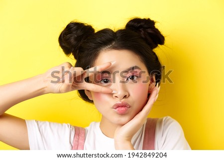 Close-up of cute asian woman with bright glamour makeup, long fingernails, showing v-sign and pouting silly at camera, standing kawaii on yellow background