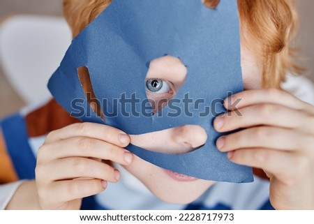 Close-up of cute blueeyed little boy covering face with handmade blue paper mask while keeping eye and nose in cut holes