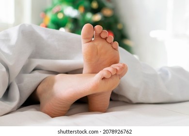 Close-up Of Cute Child Baby Feet Barefoot Covered In A Blanket, Kid Relaxing On White Bed On Background Of Christmas Tree. Cozy Merry Christmas Morning At Home, Joyful Moments In Winter Holiday Time.