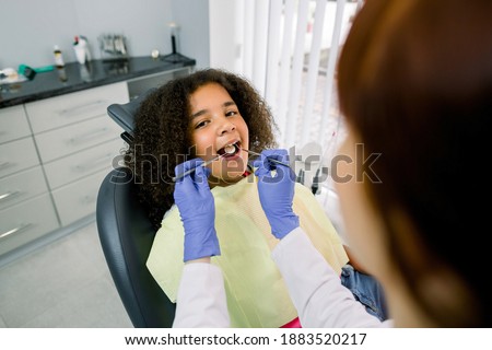 Close-up of cute curly mixed raced girl with open mouth during oral checkup at the dentist. Young female Caucasian dentist in latex gloves, making teeth examination with tools