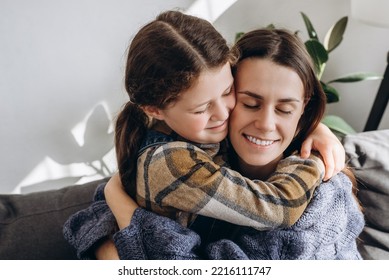 Close-up Of Cute Little Daughter And Happy Young Mom On Cheek Hugging Her From Behind, Funny Caring Child Piggyback Embrace Smiling Mother Showing Love. Kid Girl And Mommy Have Sweet Moment Together