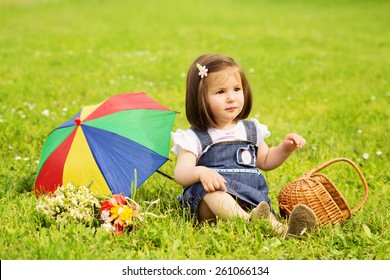 Closeup Of Cute Little Toddler Girl On Picnic In Park In Spring Sitting. Happy Beautiful One Year Old Girl With Parasol, Basket And Flowers Outdoors Portrait. Mildly Retouched, Horizontal.