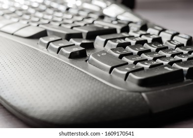 Close-up Of A Dark Dusted Computer Keyboard. Black Ergonomic Keyboard On A Light Gray Desk. Eye Level Shooting. Selective Focus.