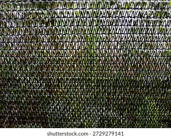 Close-up of dark mesh netting with triangular weave, revealing green foliage behind; a geometric texture forming a semi-transparent screen between viewer and garden.