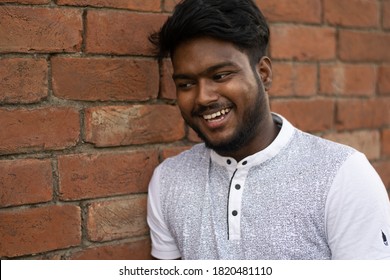 Closeup of a dark skinned young man posing beside old red brick wall. Relax profile picture with a confident smile on face | Indian dark skinned boy laughing